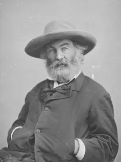 Black-and-white portrait of bearded man seated, wearing a floppy hat, with his hands in his jacket pockets.