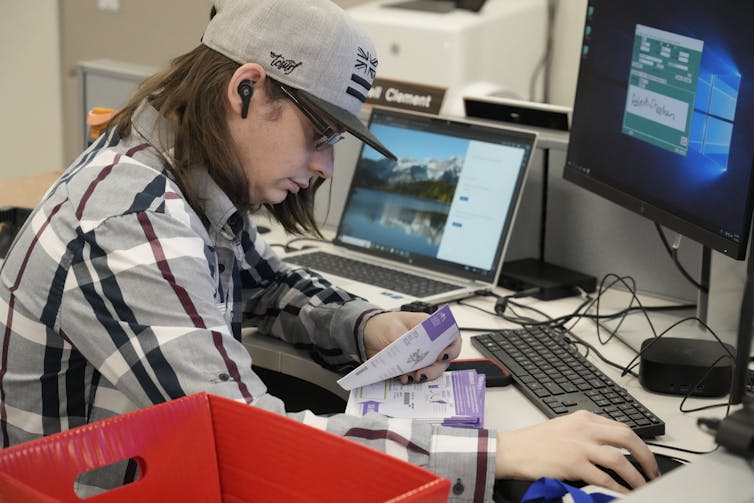 Young man in a hat sits in front of a computer holding a ballot in one hand and the computer mouse in the other