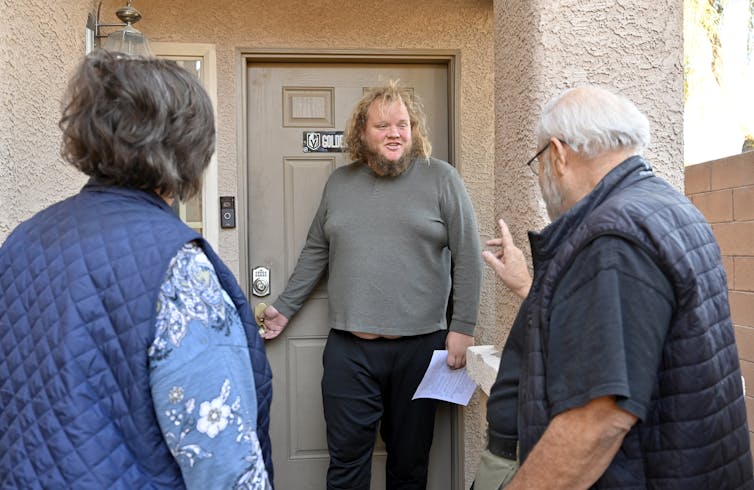 Two people speak with a man at his front door