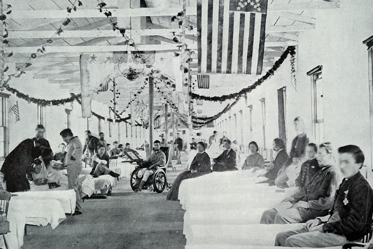Black and white photo of large room with American flags hanging from the ceiling and beds with wounded troops in them.