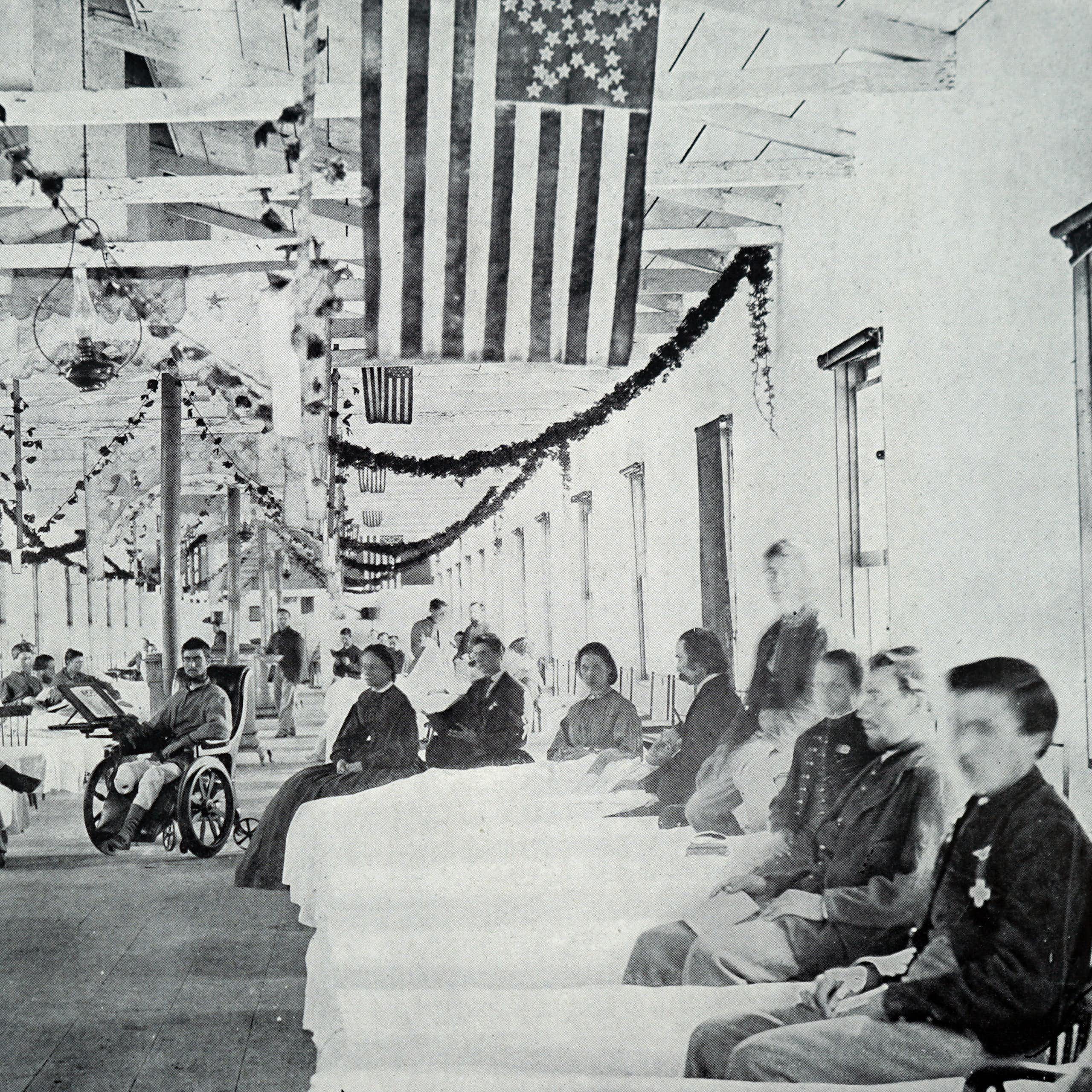 Black and white photo of large room with American flags hanging from the ceiling and beds with wounded troops in them.