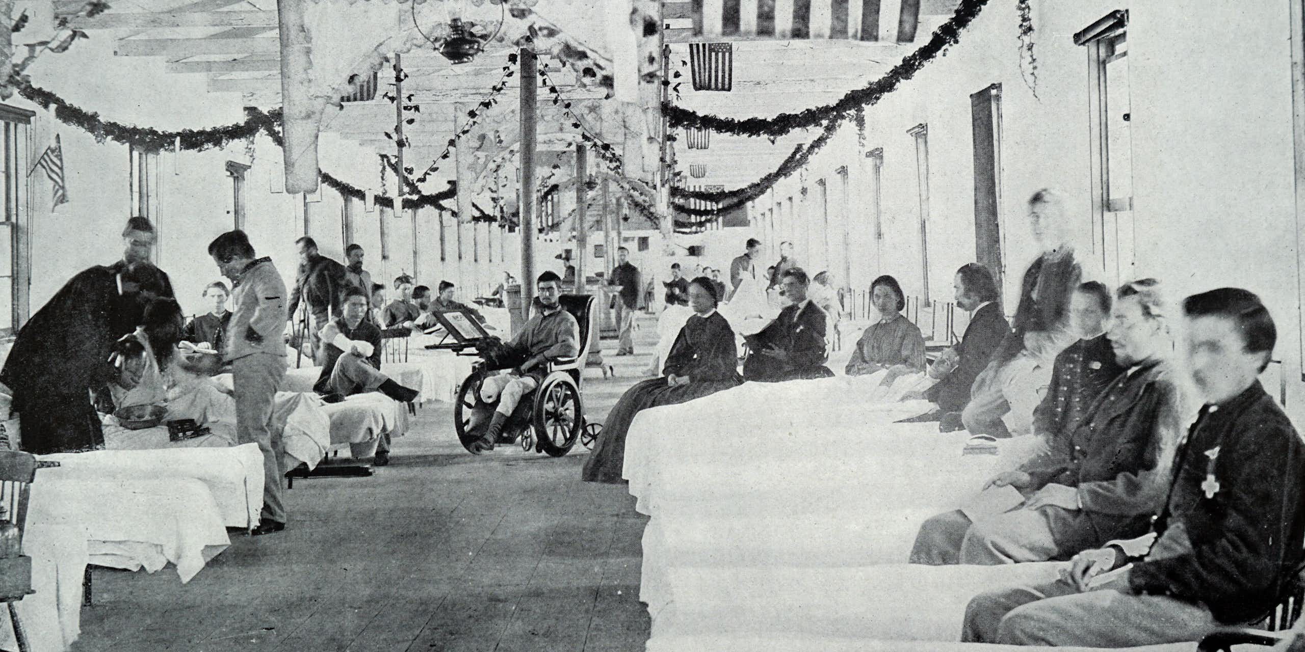 Black and white photo of large room with American flags hanging from the ceiling and beds with wounded troops in them.