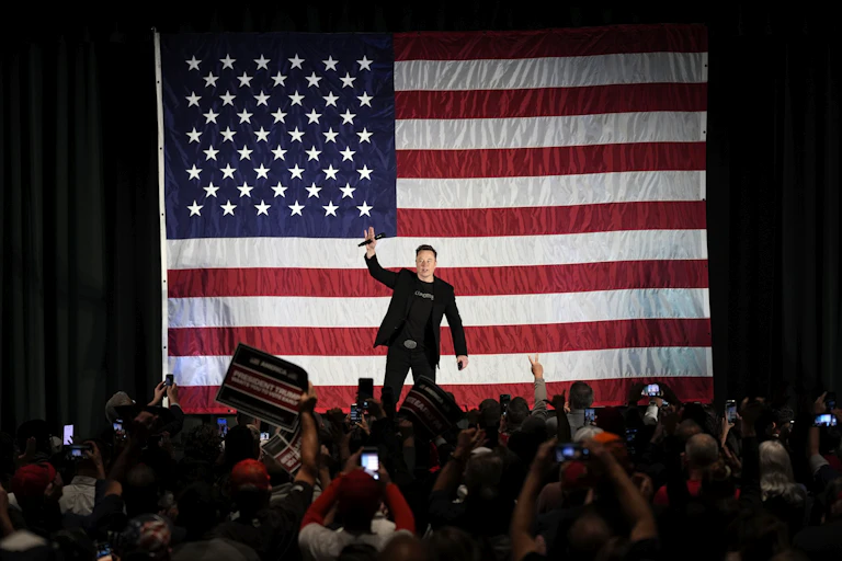 Crowd of people taking photos of a man on stage holding a microphone in front of a large American flag.