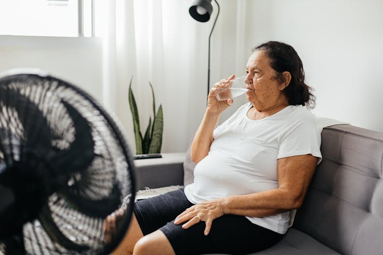A woman sitting on a couch drinking a glass of water in front of a fan.
