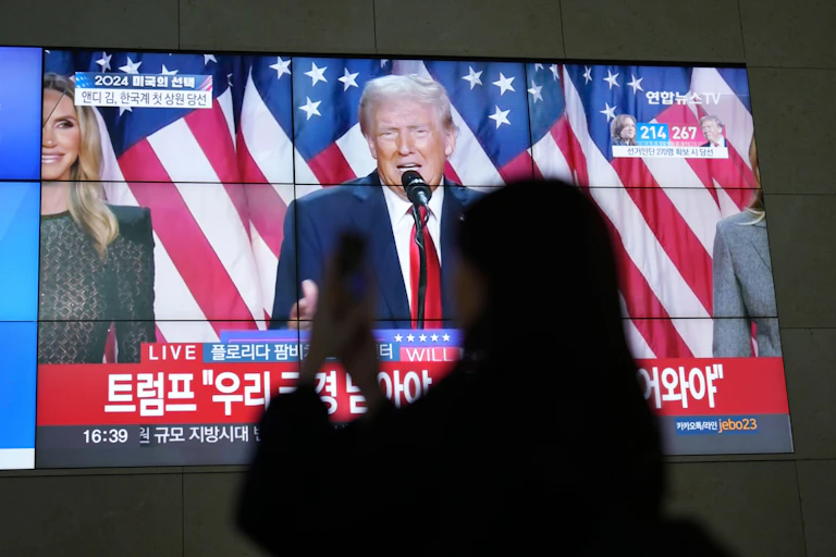 A silhouette of a woman looking at her phone with Donald Trump on a TV screen behind her.