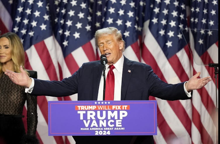 A man with blondish-grey hair stands at a lectern with his arms spread. American flags are behind him.