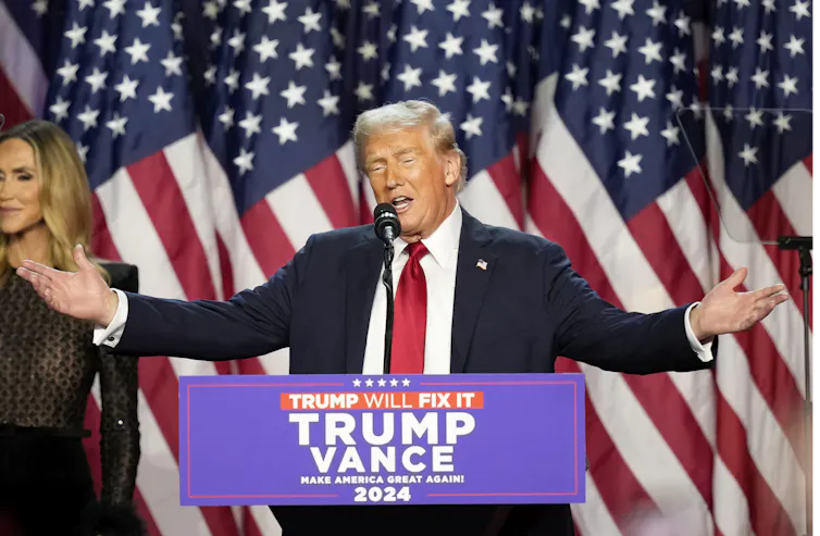 A man with blondish-grey hair stands at a lectern with his arms spread. American flags are behind him.