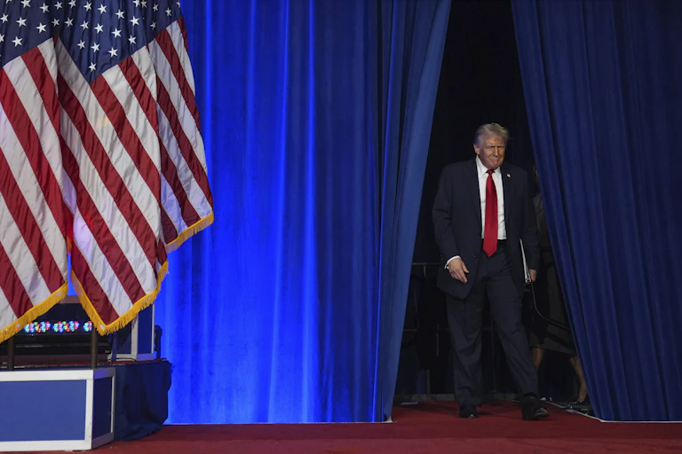 A man with blond-grey hair smiles as he walks onto a stage. A large American flag is beside him.