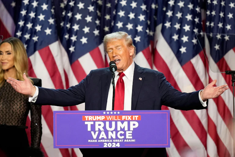 Donald Trump stands at a podium with his arms outstretched.