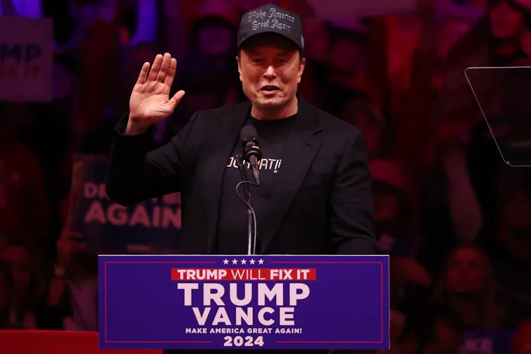 Elon Musk stands at a podium, raising his right hand.