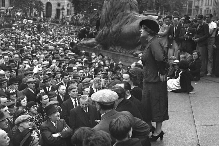 Black and white photo of Wilkinson speaking to a large crowd of mostly men, around one of the lion statue in Trafalgar Square