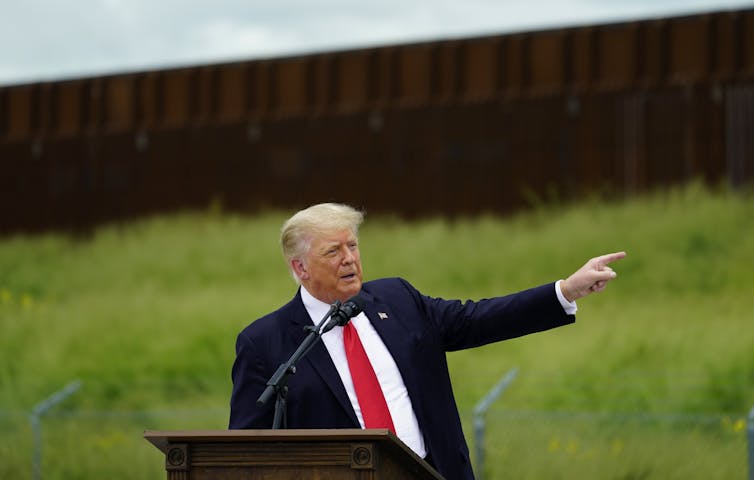 A man in a blue suit and red tie points at a large wall behind him.