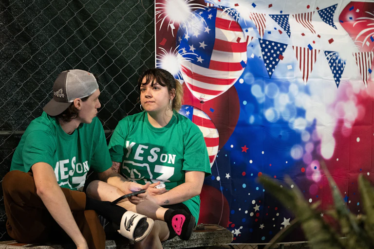 Two people wearing green T-shirts that read "Yes on 3" sit in front of a patriotic American banner