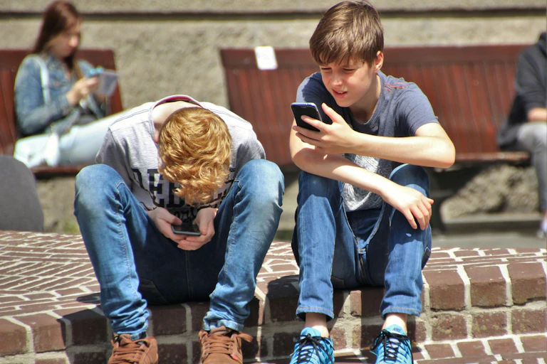 Two young boys in jeans sit on a kerb outdoors looking at their phones.