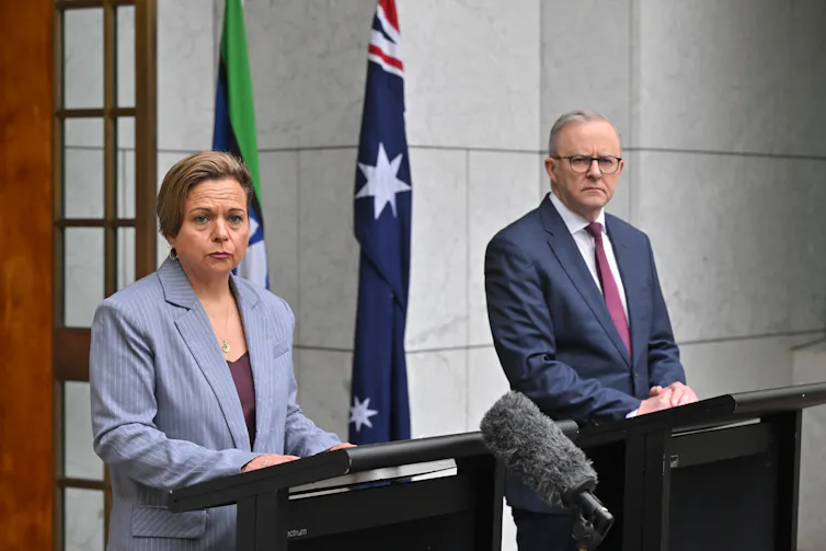 A woman in blue blazer and a man in dark suit stand at podiums at a press conference with Australian flag behind them.