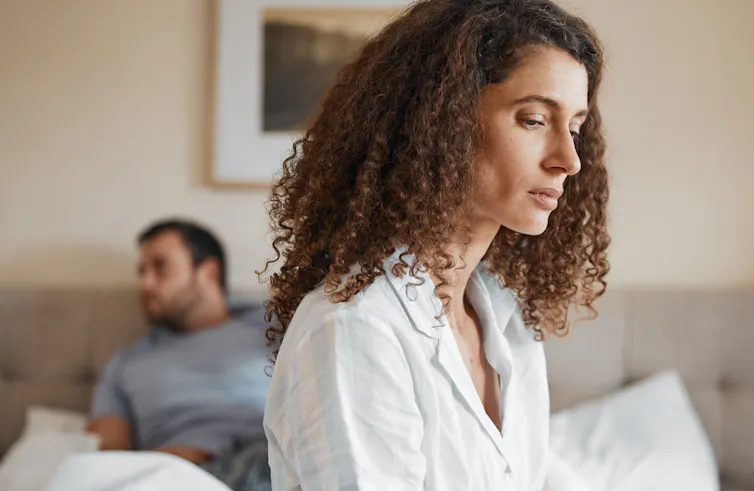 A woman sits on the edge of a bed with a pensive expression, a man siting behind her looks off into the distance