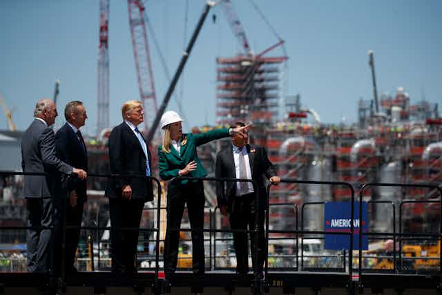 Donald Trump stands with men in suits while a woman in a hard hat points out something in the distance. construction is underway behind them.