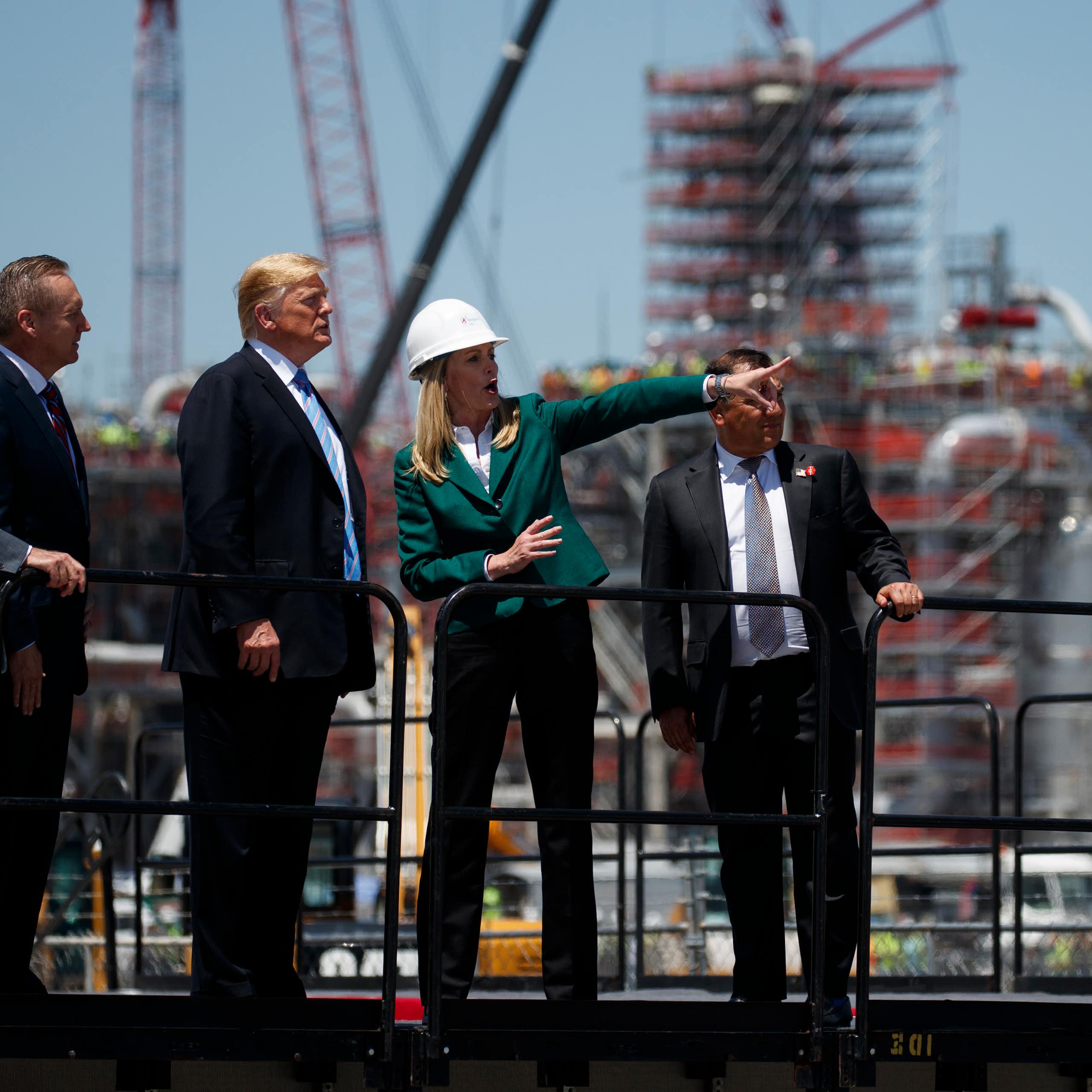 Donald Trump stands with men in suits while a woman in a hard hat points out something in the distance. construction is underway behind them.