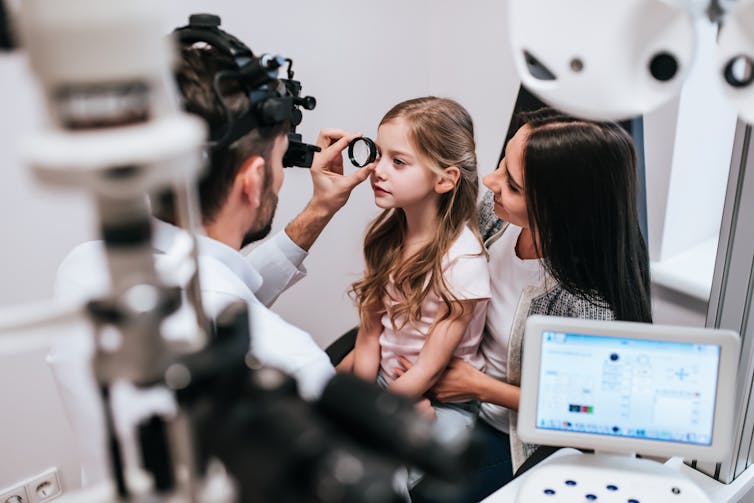 An optometrist holds a magnifying glass to the eye of a little girl as she sits on her mother's knee.
