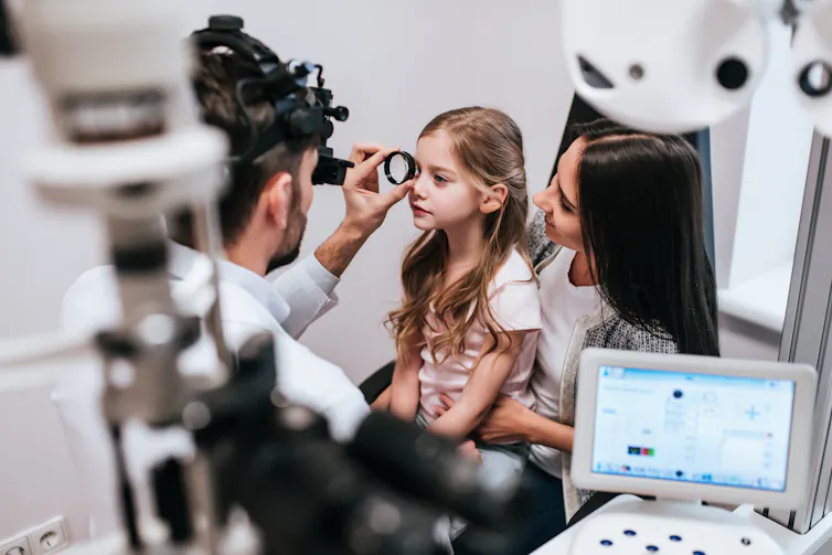 An optometrist holds a magnifying glass up to a young girl's eye as she sits on her mother's knee.