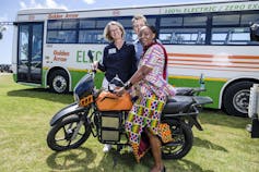 Three people stand with an orange electric motorbike in front of an electric bus