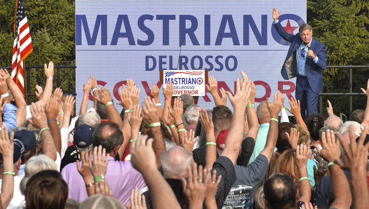 A man in a blue suit speaks to a crowd with his one hand raised. In the background is a large billboard of Mastriano Delrosso