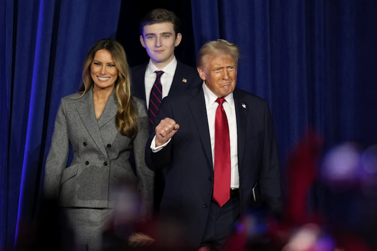 A light-haired man in a black suit and red tie, flanked by a smiling woman in a gray jacket and a tall young man, raises his fist.