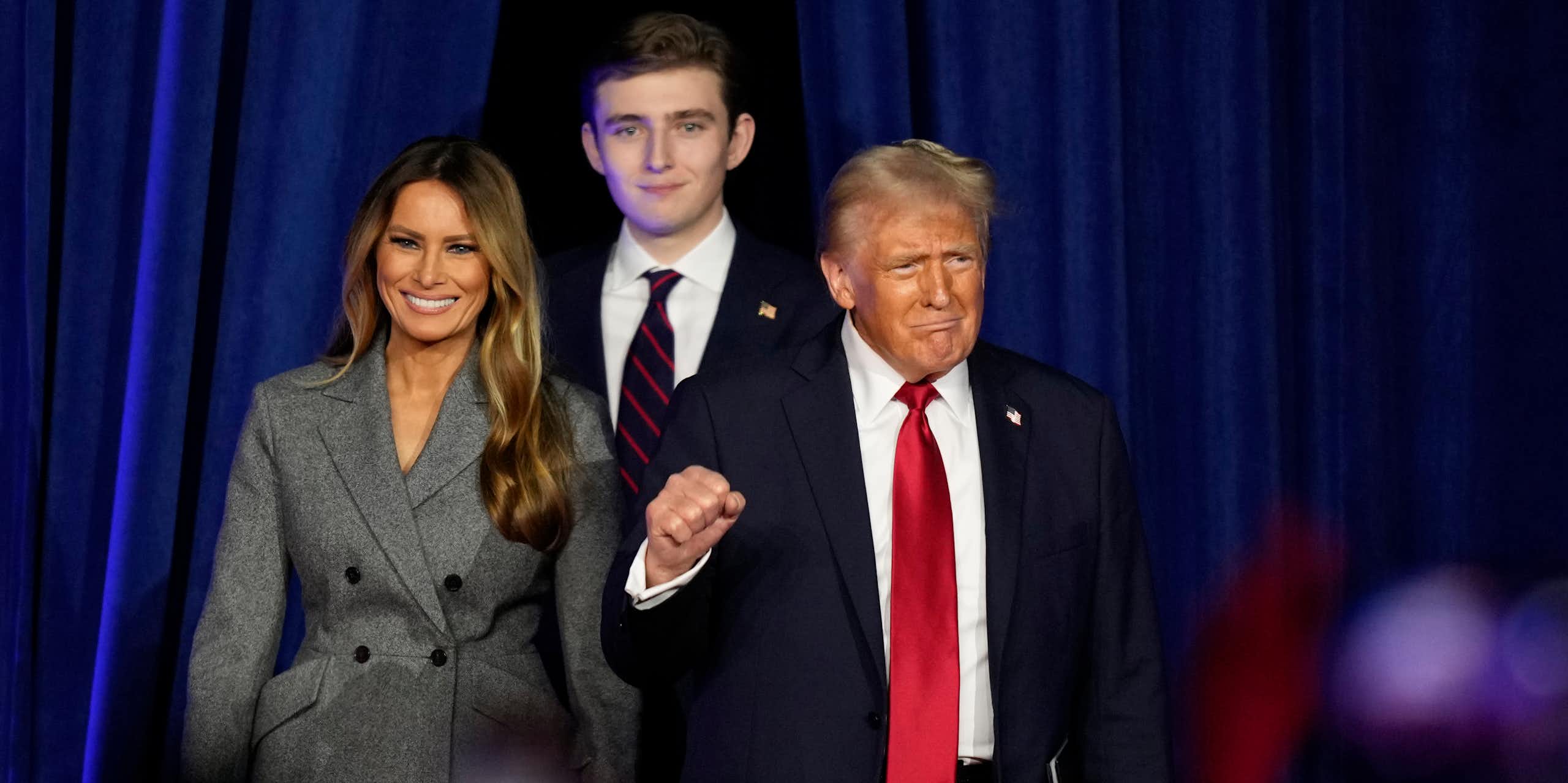 A light-haired man in a black suit and red tie, flanked by a smiling woman in a gray jacket and a tall young man, raises his fist.