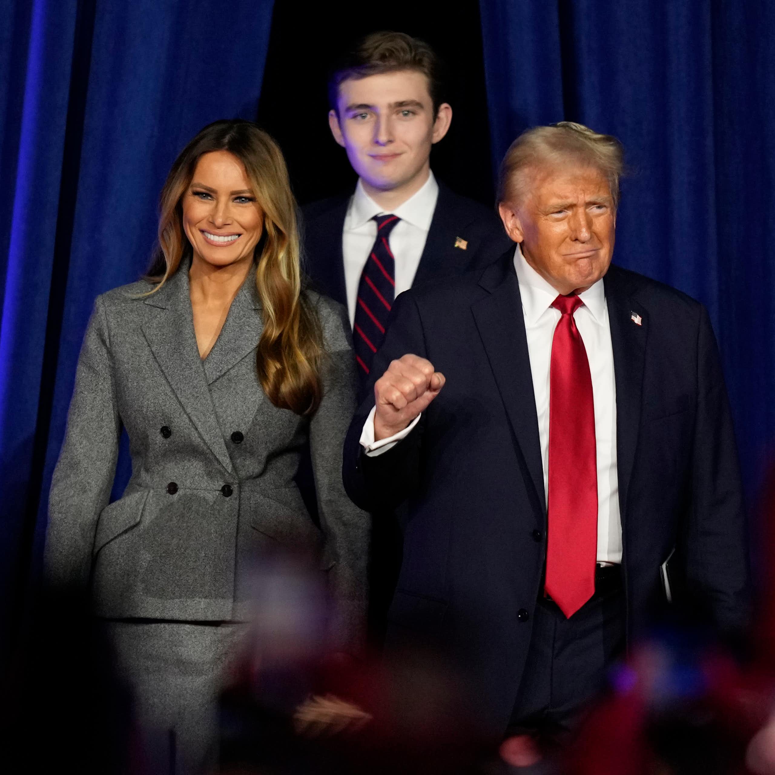 A light-haired man in a black suit and red tie, flanked by a smiling woman in a gray jacket and a tall young man, raises his fist.