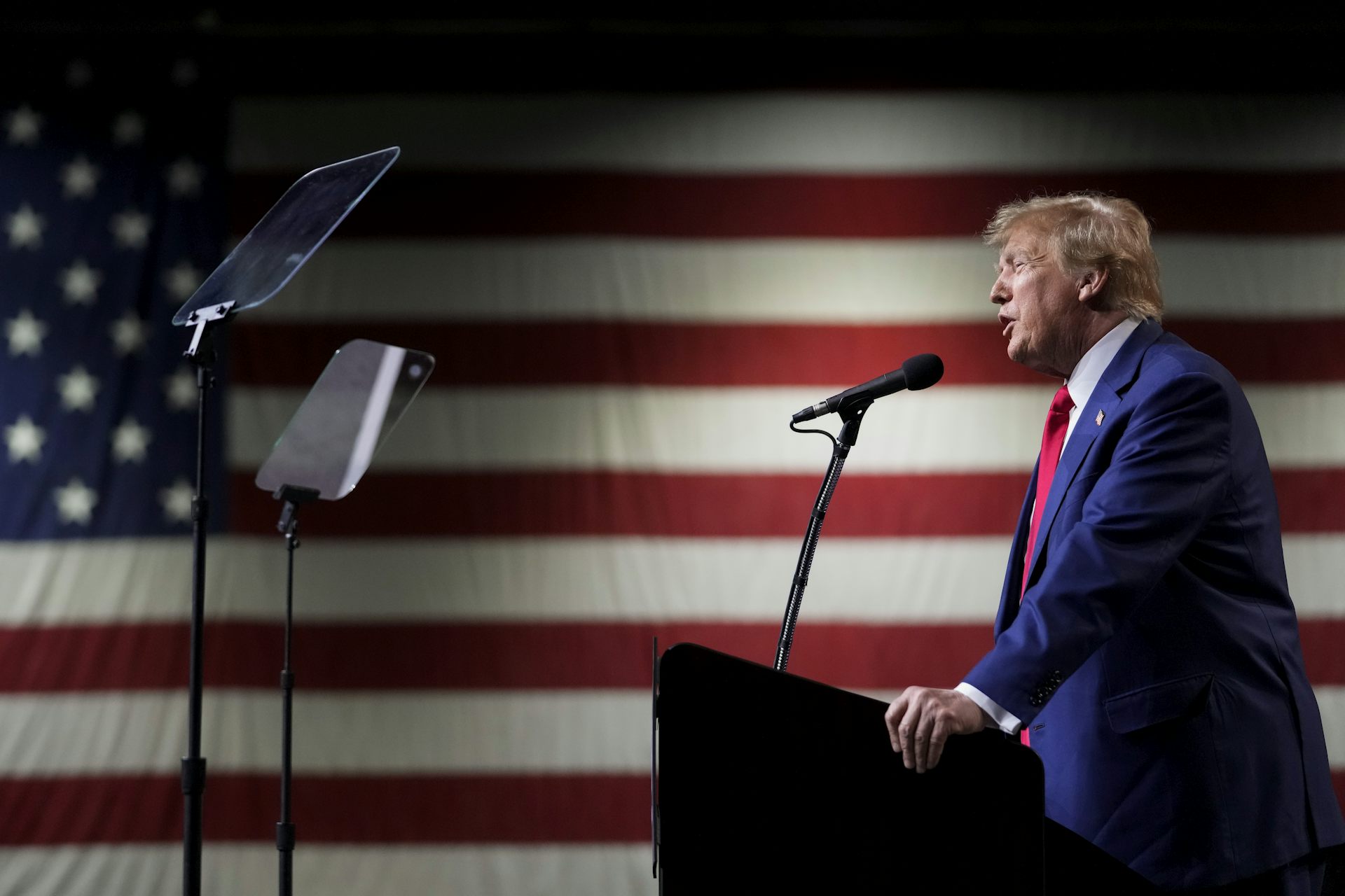 A man is photographed from the side speaking into a microphone with a large American flag as a backdrop.