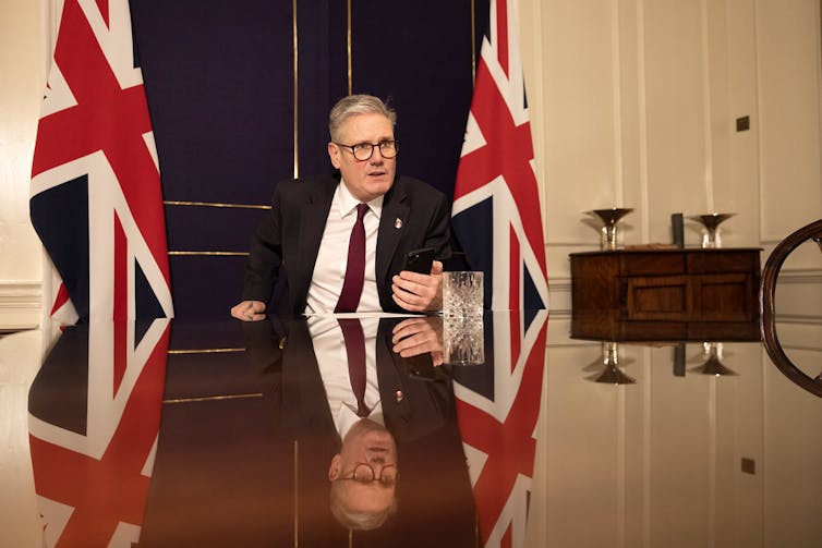 Starmer sitting at a large table, holding an iphone and speaking. Two UK flags are behind him on either side.