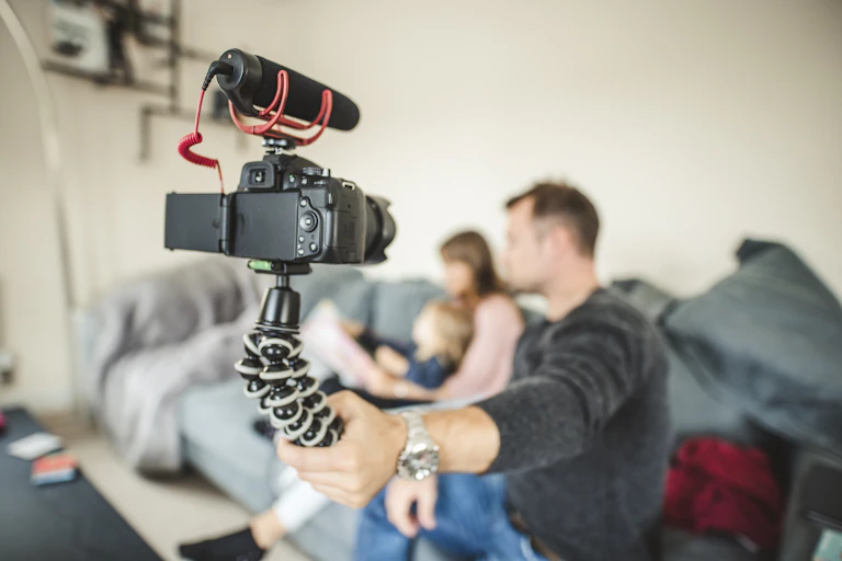 A father holds up a camera to record a mother and her young child reading a book together on a sofa.