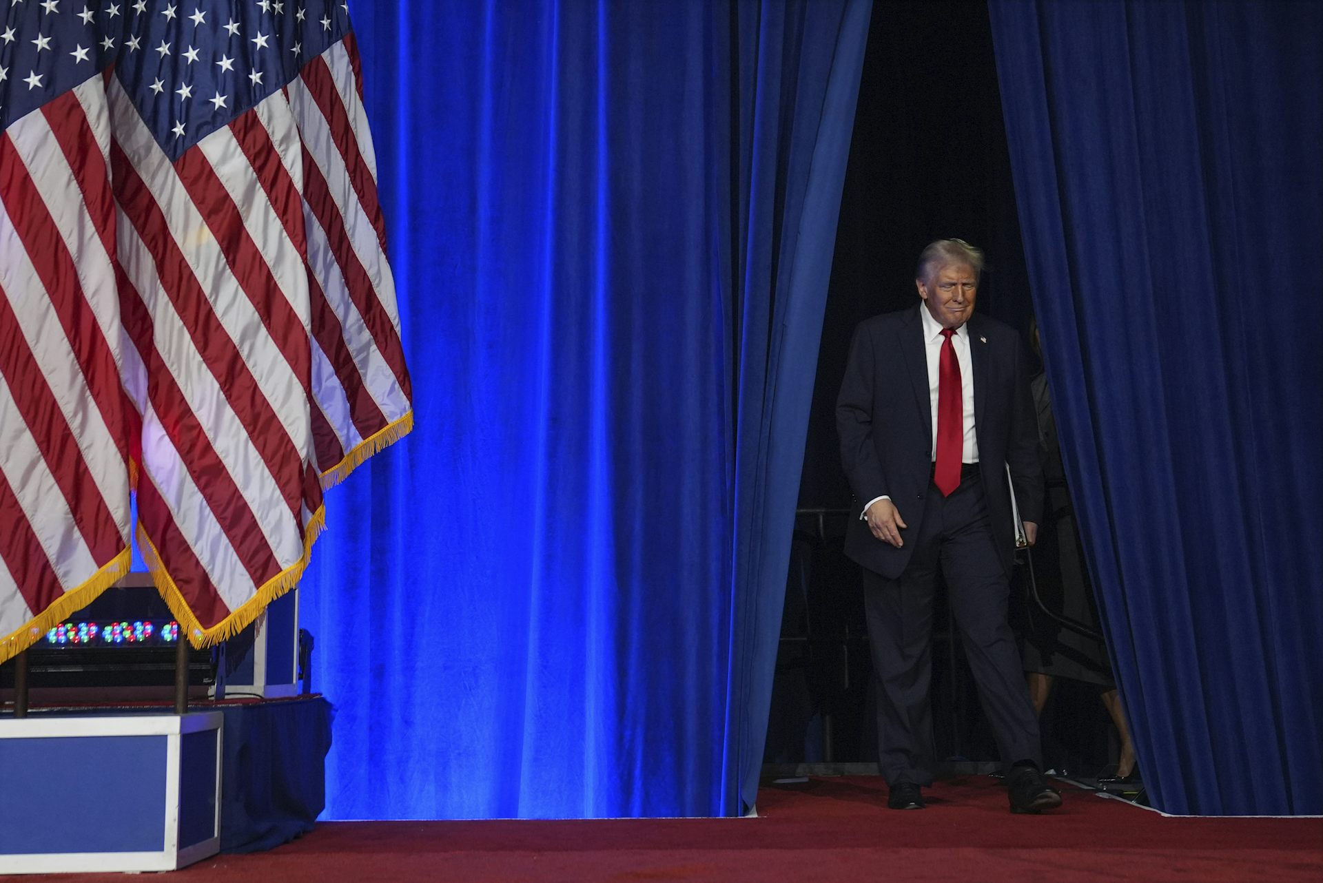 Un homme aux cheveux blond sourie en montant sur scène. Un grand drapeau américain est dressé à ses côtés.