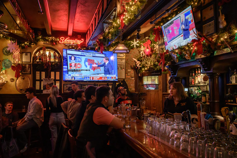 People gather in a warmly lit and festively decorated bar while election night coverage airs on TV screens
