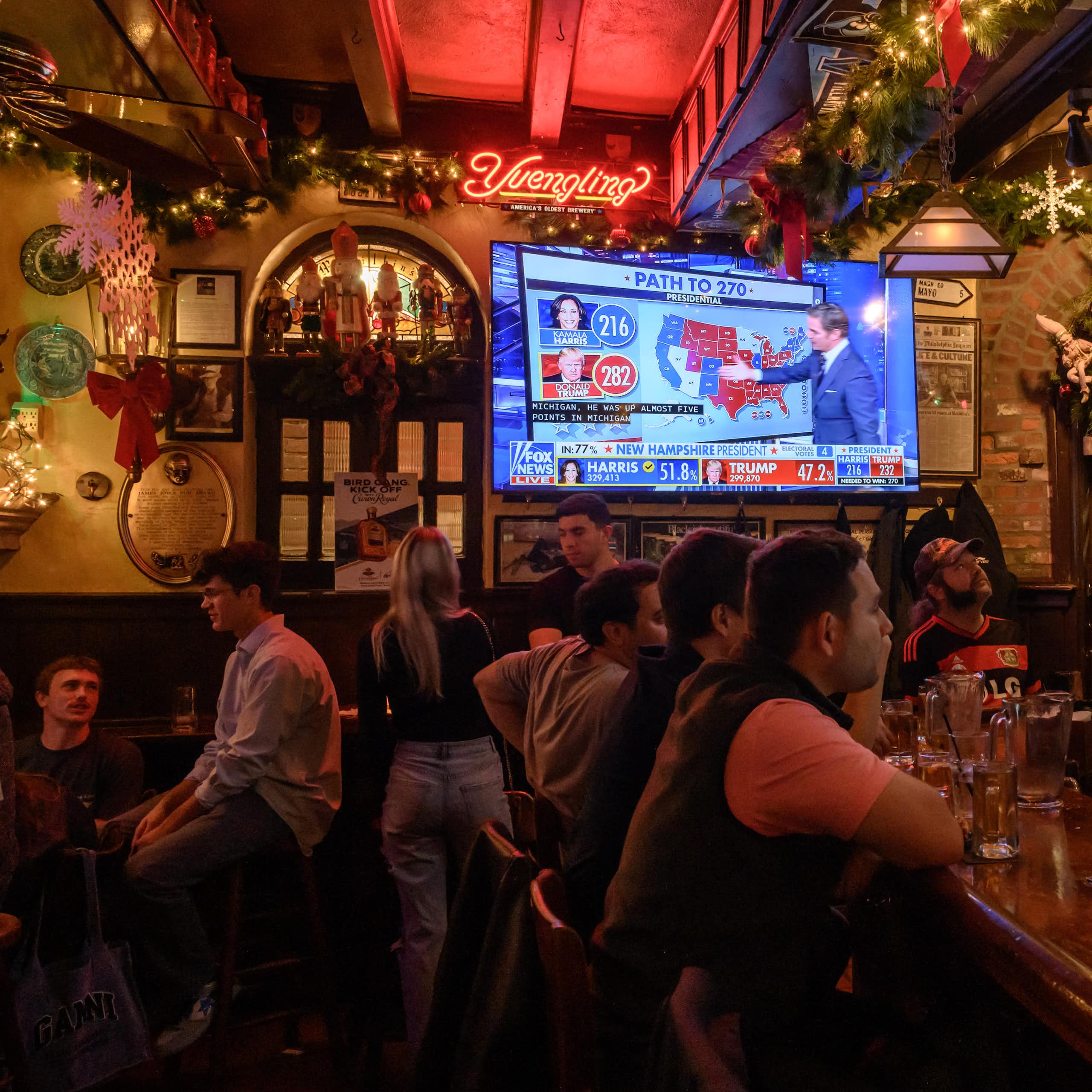 People gather in a warmly lit and festively decorated bar while election night coverage airs on TV screens