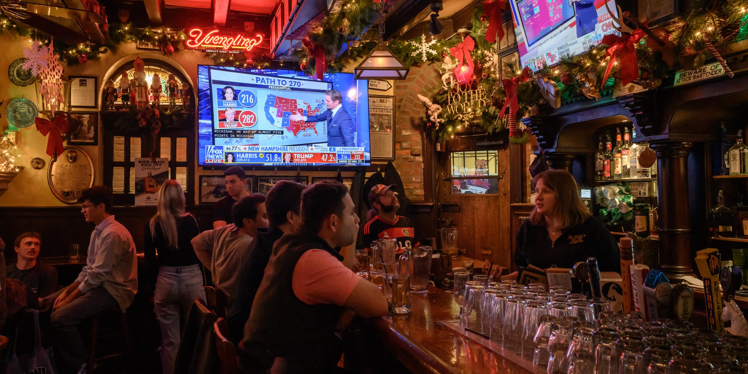 People gather in a warmly lit and festively decorated bar while election night coverage airs on TV screens