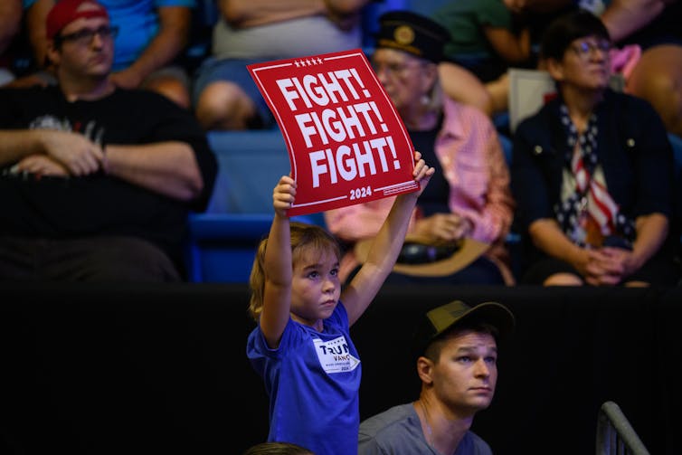 Child wearing blue Trump T-shirt holds red sign that says