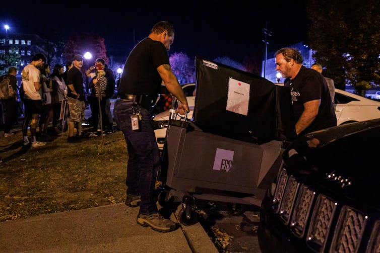 Two men in black T-shirts push a cart containing a polling machine as onlookers stand nearby in evening