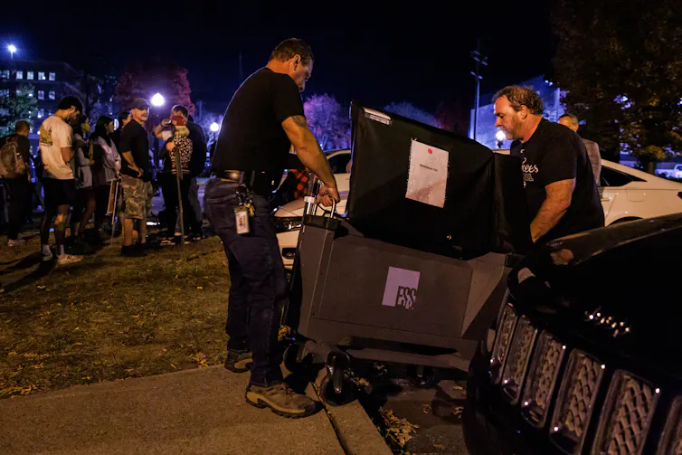 Two men in black T-shirts push a cart containing a polling machine as onlookers stand nearby in evening