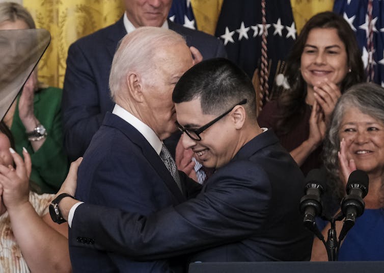 A man with glasses and a black shirt hugs an older white man with a blue suit. People behind them clap.