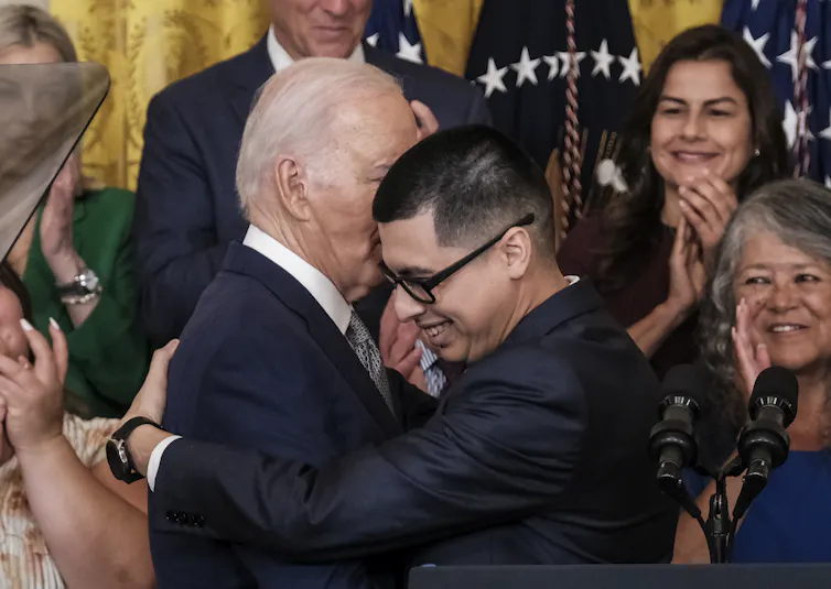 A man with glasses and a black shirt hugs an older white man with a blue suit. People behind them clap.