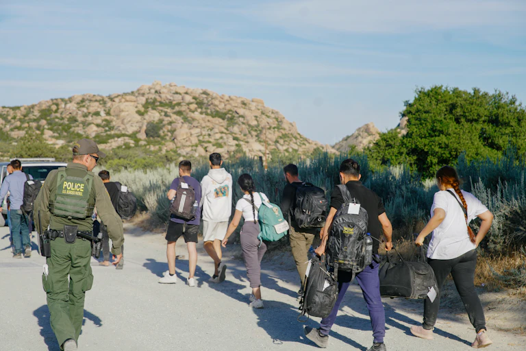 People are seen walking in a desert, with a man wearing army green and a vest that says 'police' on it in yellow watches them.