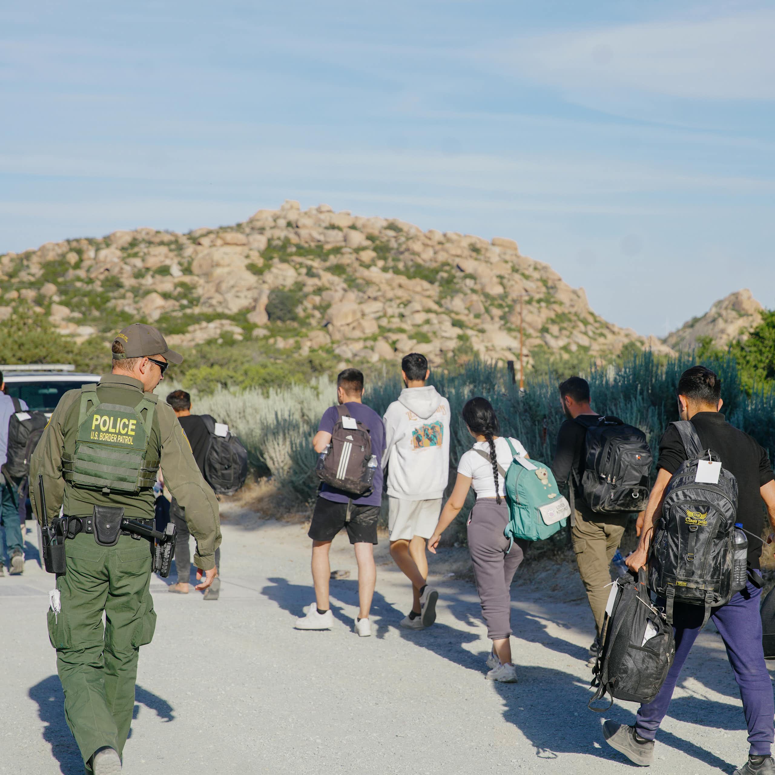 People are seen walking in a desert, with a man wearing army green and a vest that says 'police' on it in yellow watches them.