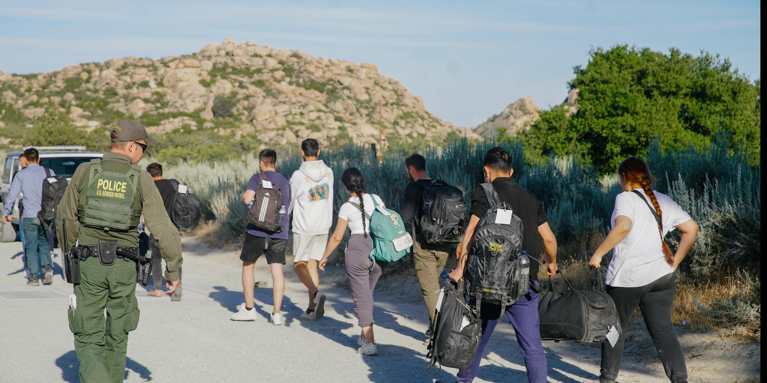 People are seen walking in a desert, with a man wearing army green and a vest that says 'police' on it in yellow watches them.