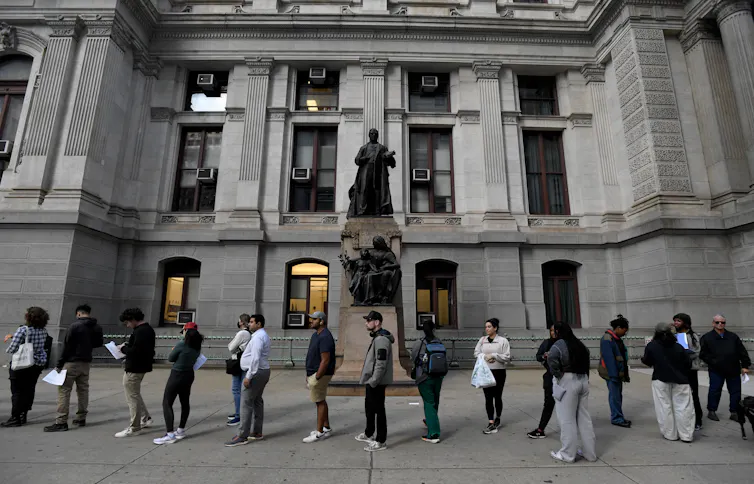 About a dozen people standing in line in front of large grey concrete building