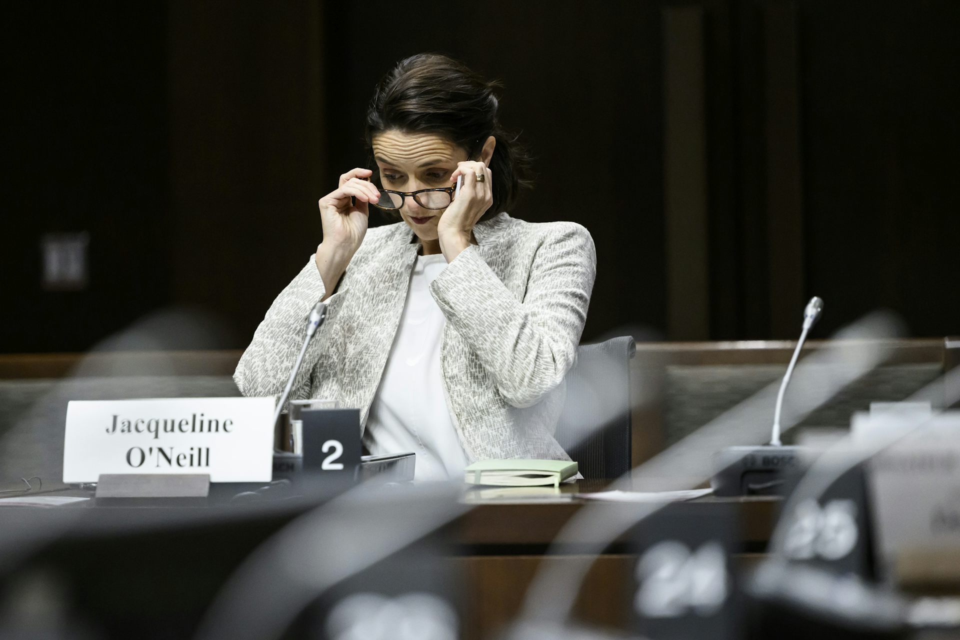 A dark-haired woman in a pale jacket puts her glasses on in a hearing room.