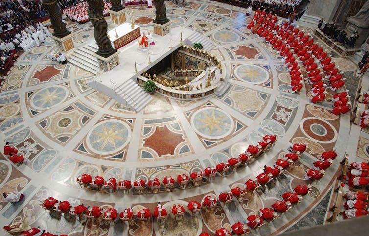 A bird's-eye view of a circular, ornate worship space, with rows of men wearing red robes.