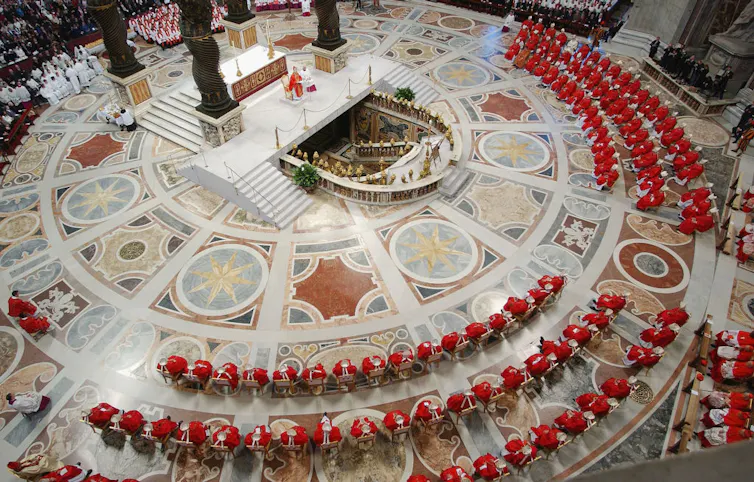A bird's-eye view of a circular, ornate worship space, with rows of men wearing red robes.