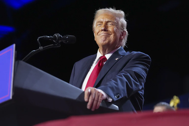 A smiling man at a lectern in a blue jacket, white shirt and red tie.