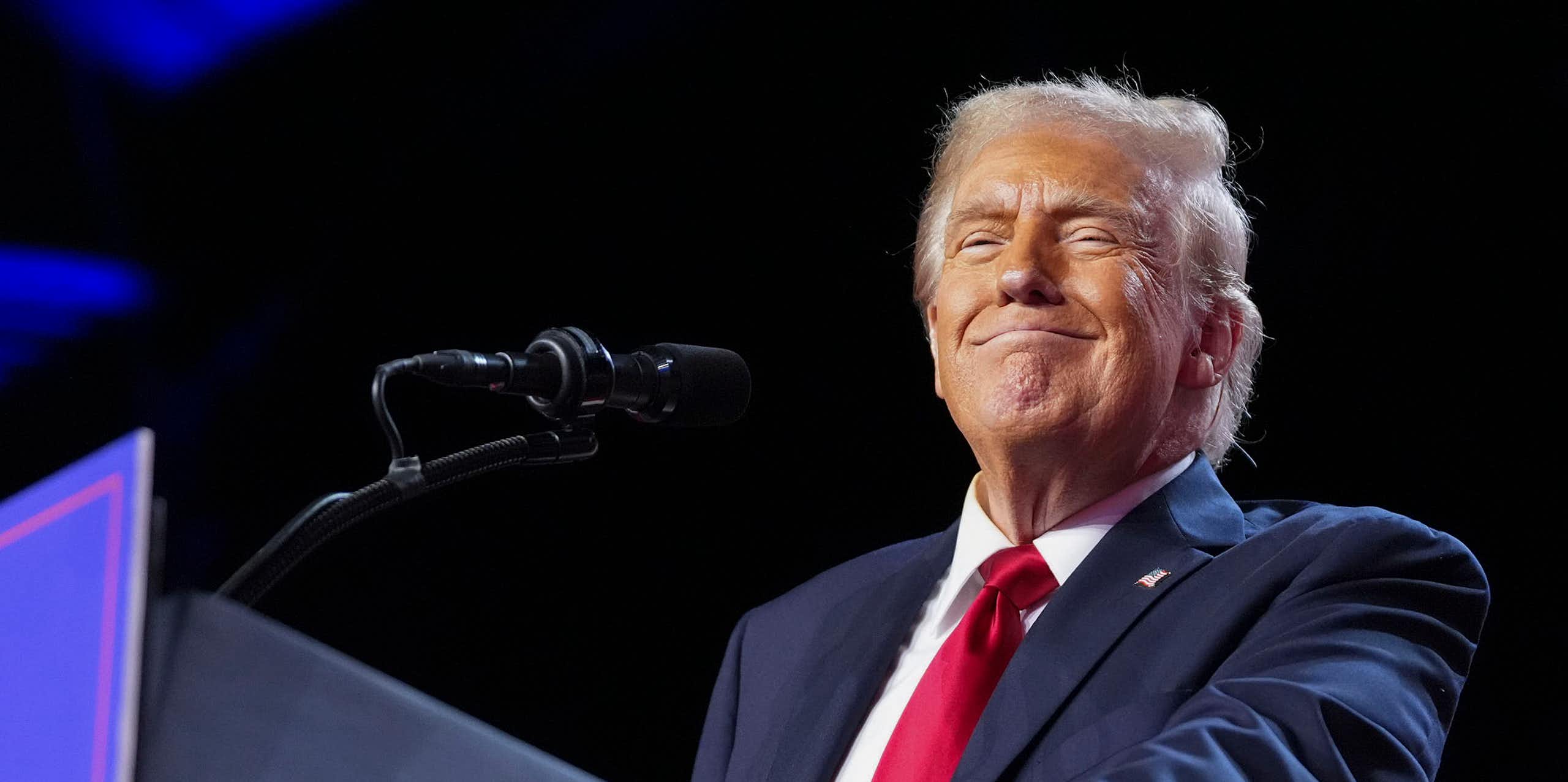 A smiling man at a lectern in a blue jacket, white shirt and red tie.